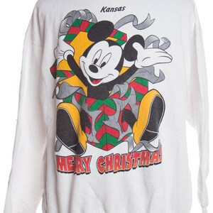 Könnte beinhalten: Weißer Langarm-Pullover mit einem Mickey-Mouse-Weihnachtsmotiv. Mickey Mouse trägt eine rote, grüne und gelbe Geschenkbox mit einem silbernen Band. Der Text "Kansas" und "Frohe Weihnachten" ist auf dem Pullover aufgedruckt.