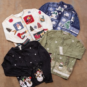 Op de afbeelding: Vier vesten met lange mouwen voorzien van kerstborduursels en applicaties. De collectie bevat een cr&egrave;mekleurige trui met sneeuwpoppen, een blauw vest met rits en sneeuwvlokken, een groen vest met wintertafereel en een zwart vest met knopen.