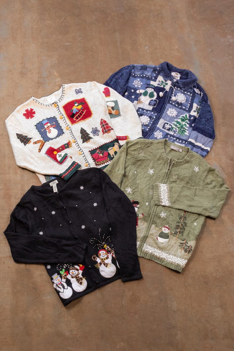 Op de afbeelding: Vier vesten met lange mouwen voorzien van kerstborduursels en applicaties. De collectie bevat een cr&egrave;mekleurige trui met sneeuwpoppen, een blauw vest met rits en sneeuwvlokken, een groen vest met wintertafereel en een zwart vest met knopen.