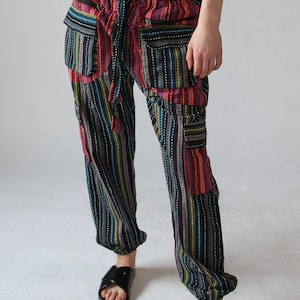 May include: Multi-coloured striped trousers with pockets. The trousers feature vertical stripes in various colours, including red, blue, green, and yellow, with a drawstring waist and elastic cuffs. The trousers are worn with black sandals.