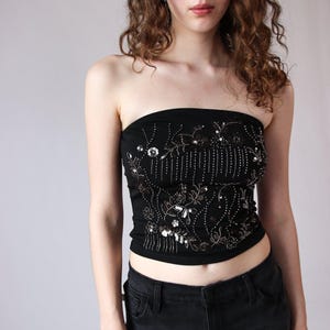 Puede incluir: Top negro sin tirantes adornado con lentejuelas y cuentas plateadas y blancas, con un diseño floral y lineal. La modelo también lleva vaqueros negros.