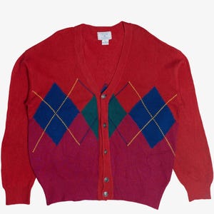 May include: Red argyle cardigan sweater with a V-neck and button closure. The sweater features a color block design with red, purple, blue, and green diamond patterns. The long-sleeved sweater is displayed on a white background.