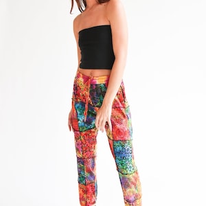 Op de afbeelding: Een persoon draagt een zwarte tube top en een kleurrijke patchwork broek. De broek heeft een levendige mix van rode, oranje, gele, groene en blauwe vierkanten met patronen. Zwarte high-top sneakers maken de outfit compleet.