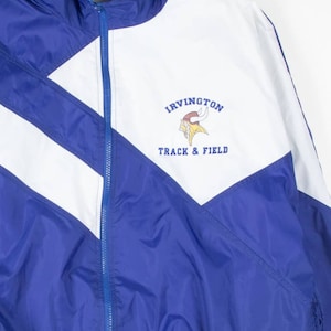 May include: A blue, white, and purple track jacket with a full zipper. The jacket features the text "IRVINGTON TRACK & FIELD" and a Viking logo. The jacket has a high collar and a diagonal color block design.
