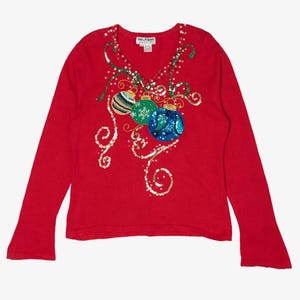 May include: Red long-sleeve Christmas sweater with a V-neck. The sweater features a design of three Christmas ornaments in blue, green, and striped patterns, embellished with sequins and gold swirls.