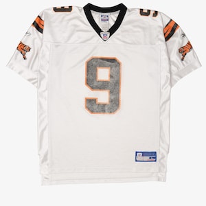May include: A white short-sleeved sports jersey with a black V-neck collar. The number 9 is printed in black with an orange outline on the front. Each sleeve features a tiger graphic and black stripes. An NFL logo is at the neck and a tag is at the hem.