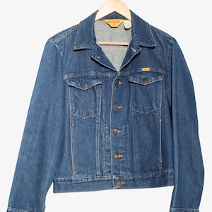May include: A blue denim jacket with a classic design, featuring a button-up front, two chest pockets, and two side pockets. The jacket is hanging on a wooden hanger. The brand name "Hustler" is visible on a tag.