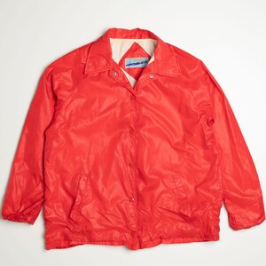 May include: A red nylon jacket with a button closure and a white label on the collar. The jacket has a pocket on the left side.