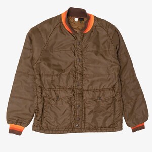 May include: A brown quilted jacket featuring a snap-button front closure, two front flap pockets, and a ribbed collar and cuffs with orange and red stripes. The jacket has long sleeves and a horizontal quilted pattern throughout the fabric.