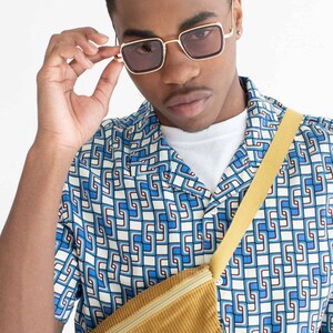 Może przedstawiać: Osoba nosząca okulary przeciwsłoneczne w złotej oprawie z brązowymi soczewkami, koszulę zapinaną na guziki we wzór w kolorze niebiesko-białym, biały podkoszulek i musztardową nerkę z sztruksu.