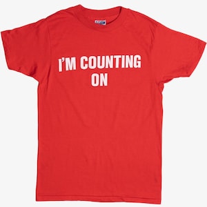 May include: Red short-sleeved t-shirt with white text that reads "I'M COUNTING ON".