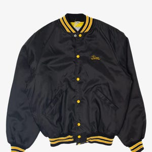 May include: Black satin bomber jacket with yellow and black striped trim on the cuffs, collar, and waist. The jacket has yellow button closures and the name "Jim" embroidered in yellow script on the chest.