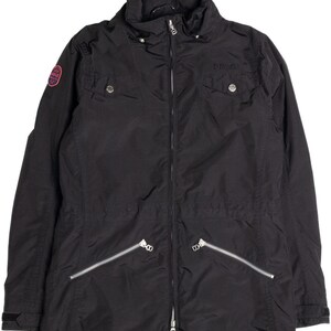 May include: Black jacket with a high collar, a zipper closure, and multiple pockets. The jacket has a small logo patch on the left sleeve and a logo above the right chest pocket. The jacket is made of a lightweight material.