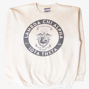 Pode incluir: Um moletom bege claro com gola redonda e um emblema circular. O emblema apresenta o texto "Lambda Chi Alpha Iota Theta" e um design de brasão. O moletom é feito de um material macio e tem um ajuste clássico.