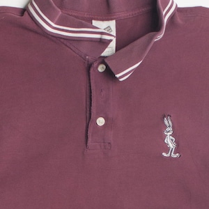 May include: Maroon polo shirt with a white and light blue striped collar. The shirt features a Bugs Bunny embroidered graphic on the lower right side. The shirt has a three-button placket.
