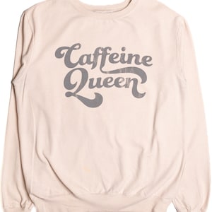 May include: A light beige sweatshirt with the text "Caffeine Queen" in a gray, retro font. The long-sleeved top has a crew neck and ribbed cuffs and hem. The casual style is suitable for everyday wear.