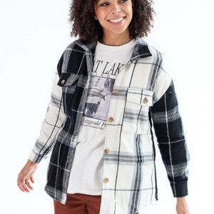 Puede incluir: Una camisa de franela a cuadros blancos y negros con una camiseta blanca debajo. La camiseta tiene un estampado gráfico de un barco y el texto "Great Lakes" y "Fitzgerald Park from Cleveland, Ohio". La persona lleva pantalones de pana marrones.