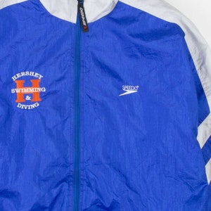May include: A blue, white, and gray Speedo track jacket. The jacket has a full zipper and features the text "Hershey Swimming & Diving" in orange and white. The Speedo logo is on the chest.