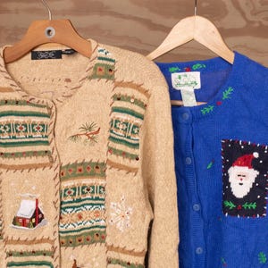 Op de afbeelding: Een beige gebreid vest met geborduurde wintertaferelen met een hut, hert en sneeuwvlokken, naast een blauw gebreid vest met knoopsluiting en een geborduurde kerstman-patch, beide hangend aan houten kledinghangers.
