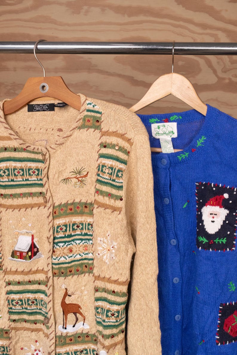Op de afbeelding: Een beige gebreid vest met geborduurde wintertaferelen met een hut, hert en sneeuwvlokken, naast een blauw gebreid vest met knoopsluiting en een geborduurde kerstman-patch, beide hangend aan houten kledinghangers.