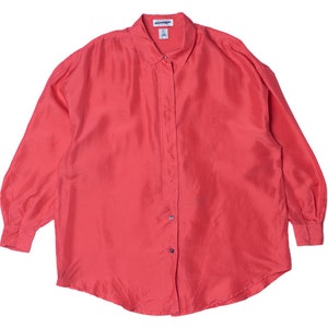Puede incluir: Una camisa con botones de color coral con mangas largas. La camisa tiene un cuello clásico y un ajuste ligeramente holgado. La tela parece ser ligera, posiblemente seda o una mezcla de seda. La camisa está tendida sobre una superficie blanca.
