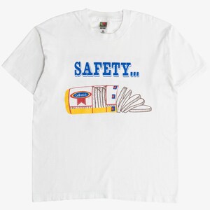 Può includere: T-shirt bianca con la scritta blu "SAFETY..." sopra un'immagine a cartoni animati di pane a fette. Il pane è in una confezione gialla e rossa con la scritta "Colonial".