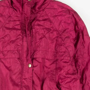 May include: A close-up of a magenta-colored jacket with a high collar and gold-colored buttons. The jacket has a quilted texture and two front pockets. The fabric has a slight sheen.