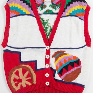 Op de afbeelding: Een rood, wit en bruin gebreid vest met een kleurrijk geometrisch patroon en een knoopsluiting. Het vest heeft een label met de tekst "Andina".