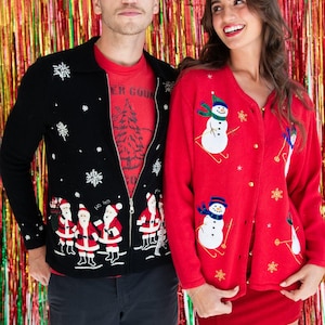 Op de afbeelding: Een zwart vest met rits, voorzien van een kerstmanpatroon en witte sneeuwvlokken, gedragen over een rood t-shirt, naast een rood vest met knopen, geborduurde sneeuwpoppen op ski's en gouden sneeuwvlokaccenten.