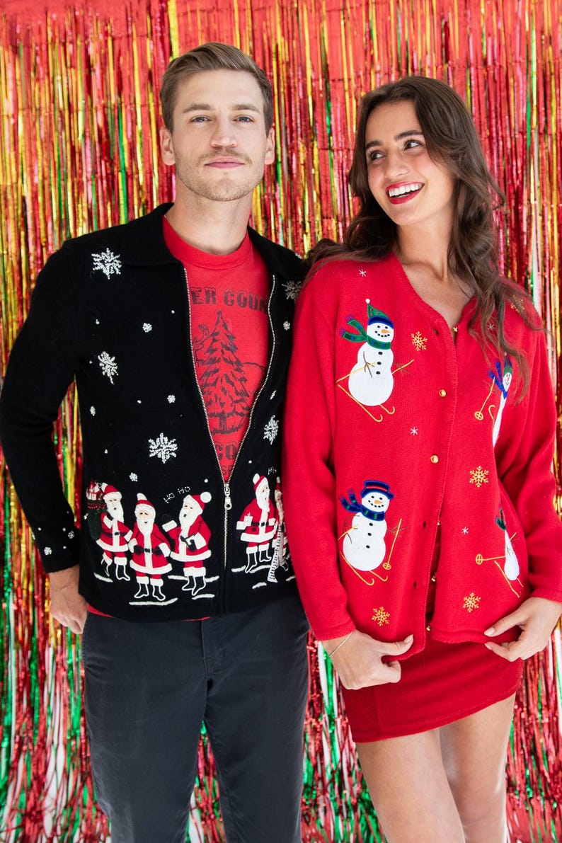 Op de afbeelding: Een zwart vest met rits, voorzien van een kerstmanpatroon en witte sneeuwvlokken, gedragen over een rood t-shirt, naast een rood vest met knopen, geborduurde sneeuwpoppen op ski's en gouden sneeuwvlokaccenten.