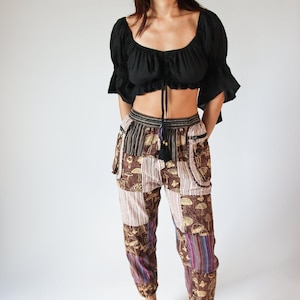 May include: A black crop top with ruffled sleeves and a drawstring neckline paired with patchwork pants. The pants feature a mix of brown, purple, and beige fabrics with mushroom and stripe patterns. Black flats complete the outfit.