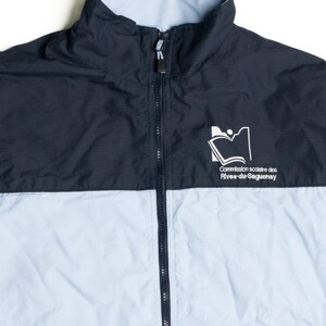 Puede incluir: Chaqueta con cremallera azul marino y celeste con cuello alto. La chaqueta presenta un logotipo blanco y texto en el pecho. La chaqueta es de dos tonos, con azul marino en la parte superior y celeste en la inferior.