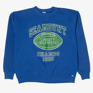 May include: Blue crewneck sweatshirt with the text "SEAMOUNT" above a green football graphic that reads "LIBERTY PATRIOTS". Below the graphic is "CHAMPS 1988" in green lettering. The sweatshirt has a small white tag on the bottom.