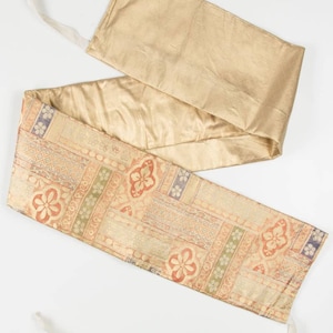 Puede incluir: Una faja larga y estrecha de tela de color dorado con un diseño estampado. La faja tiene dos lazos blancos en cada extremo.