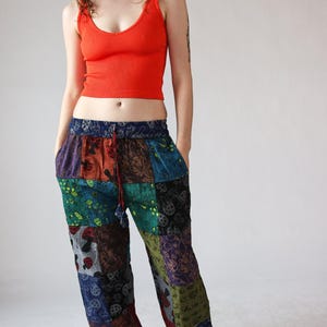 Op de afbeelding: Een oranje geribbelde tanktop gecombineerd met een patchwork broek. De broek heeft verschillende gekleurde stofvierkanten met paddenstoel- en bloempatronen. De broek heeft een taille met trekkoord en zakken. Zwarte sandalen maken de outfit af.