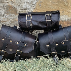 Può includere: Tre selle da moto in pelle nera con fibbie e cinghie. Le borse sono disposte a forma di piramide su un letto di fieno.