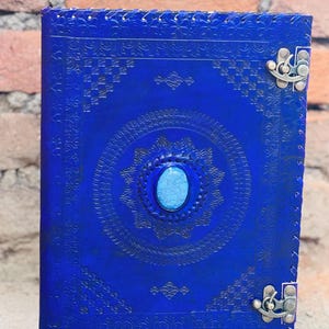 Op de afbeelding: Een kobaltblauw leren notitieboek met reliëf bloempatronen en een ovale turkooizen steen in het midden. Het notitieboek heeft metalen sluitingen en gestikte randen. De omslag heeft een gedetailleerd, symmetrisch ontwerp.