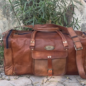 Könnte beinhalten: Eine braune Ledertasche mit einer großen Fronttasche und einem Schnallenverschluss. Die Tasche hat einen langen Schultergurt und einen oberen Griff.
