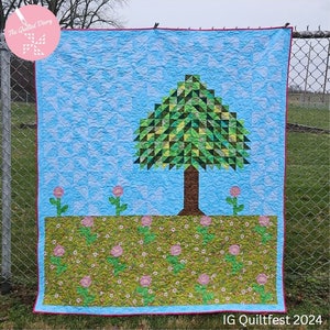 May include: A blue and green quilt with a tree in the center and a field of flowers at the bottom. The quilt is hanging on a chain link fence. The text "IG Quiltfest 2024" is at the bottom of the quilt.