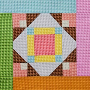May include: A colorful quilt block with a textured surface. The design features a central diamond shape with a pink square, surrounded by yellow, blue, and white geometric shapes. Brown triangles and pink squares frame the central design, set against a backdrop of green, orange, pink, and blue.