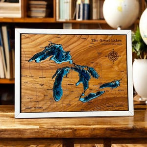 May include: A framed wooden map of the Great Lakes, with a white border. The map features a detailed, layered design showing the lakes in shades of blue against a light brown wood grain background. Text includes "The Great Lakes" and a compass rose.