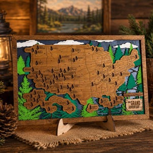 Op de afbeelding: Houten kaart van de Verenigde Staten met een gedetailleerd ontwerp. De kaart heeft een natuurlijke houten afwerking met groene en blauwe accenten en bevat kleine boomillustraties. De woorden "Grand Adventure" staan op een klein bord.