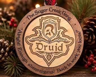 May include: Wooden Christmas ornament with the text "The Sugar Creek Gang" and "Druid" engraved. The ornament has a decorative design and a red and black plaid ribbon for hanging. The background includes pine cones and berries.