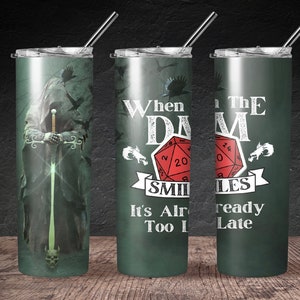 May include: A set of three stainless steel tumblers with straws. The tumblers feature a dark green background with fantasy-themed graphics. One tumbler depicts a cloaked figure with a sword, while the others display dice and text that reads "When the DM Smiles, It's Already Too Late."