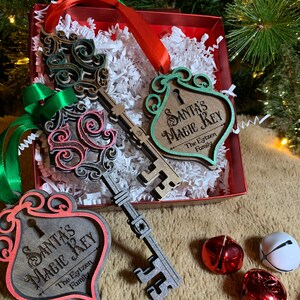 May include: Christmas ornaments in a red box with white paper filler. The ornaments are wooden keys and key-shaped tags with the text "Santa's Magic Key". The keys are in shades of gray, green, and pink. Red and green ribbons are attached.