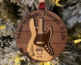 Może przedstawiać: Okrągła drewniana ozdoba świąteczna z motywem gitary. Ozdoba zawiera napis "Music Is Magic, Therapy, & Life" oraz imię "Matthew". Gitara jest w kolorze jasnobrązowym i ciemnobrązowym.