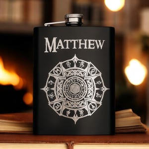 Puede incluir: Petaca negra con el nombre "MATTHEW" en letras plateadas. La petaca presenta un diseño plateado detallado con símbolos geométricos y de fantasía. La petaca tiene una tapa plateada y está sobre una superficie marrón.