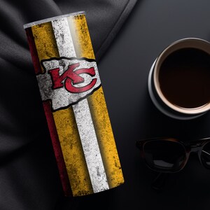 May include: A stainless steel tumbler with a white, red, and yellow design featuring the Kansas City Chiefs logo. The tumbler has a distressed look and is on a black surface.