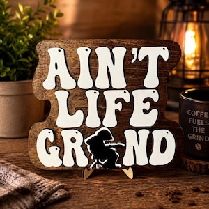 May include: Wooden sign with the phrase "Ain't Life Grand" in white, stylized letters. A black silhouette of a musician playing a guitar is featured below the text. The sign is propped up on a wooden stand.