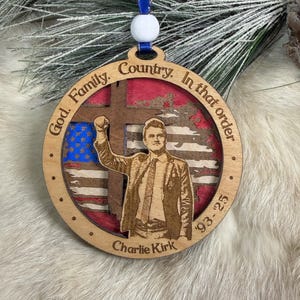May include: Wooden Christmas ornament with a blue ribbon and white bead. The ornament features a cutout design with an American flag, a man raising his fist, and the text "God. Family. Country. In that order. Charlie Kirk '93-25."